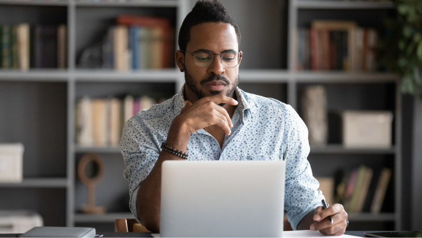 Man with glasses sits at a desk with a laptop and is thinking, bookshelf in the background.