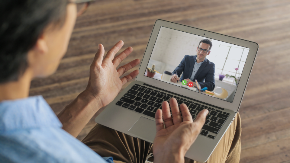 En person har videosamtal via laptop med någon som sitter vid ett skrivbord i ett ljust rum.