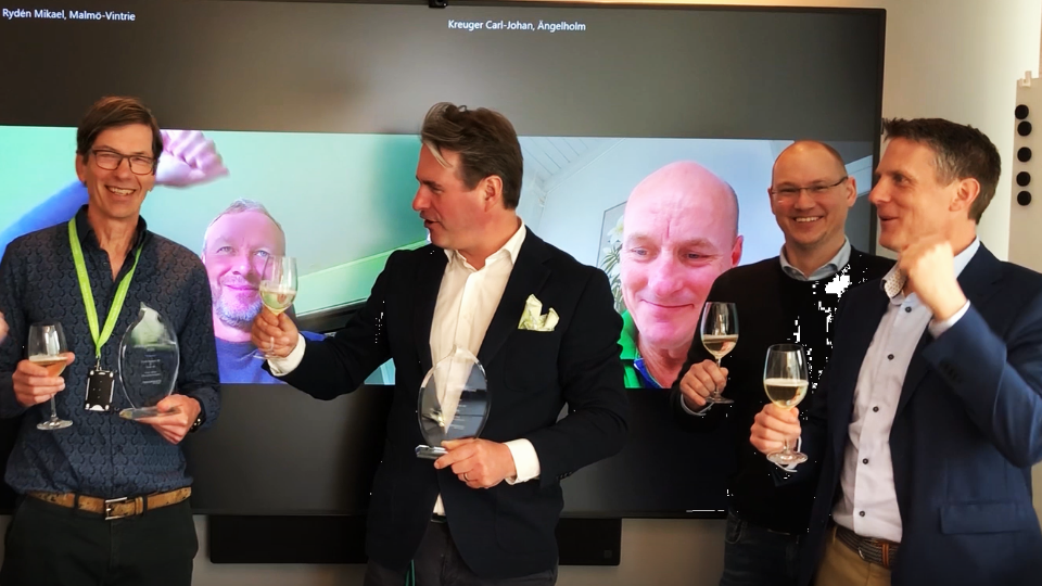 Fem män firar med glas och priser, två är på videoskärm i bakgrunden, tre står framför skärmen och ler.