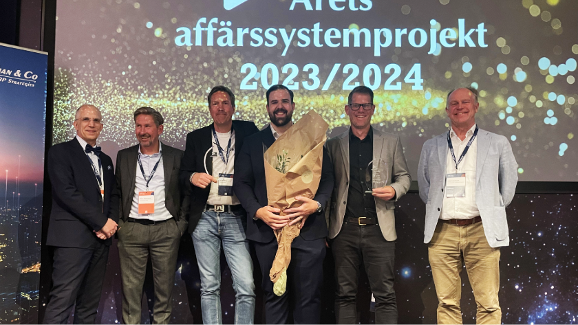 Sex män i kostym poserar på scen med blommor och pokal vid en prisutdelning, skylten visar "Årets affärssystemprojekt".