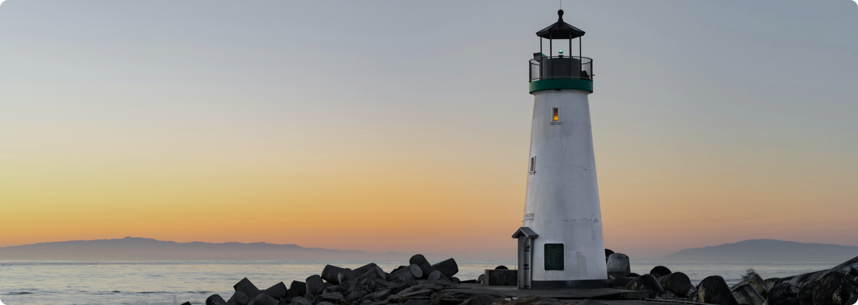 A white lighthouse stands on rocks by the sea at sunset, with the mountains faintly visible in the background.