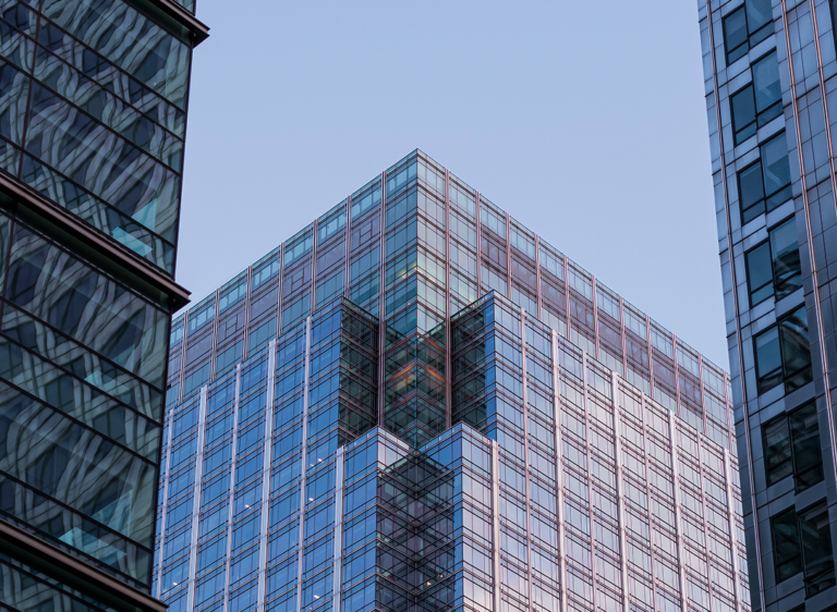 Corner of a modern glass building against a blue sky, surrounded by other high-rise buildings with glass façades.