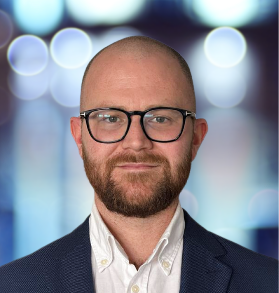 Bearded man with glasses and blazer in front of a blurred light background.