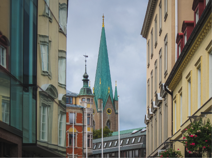 Church tower with a green pointed roof visible between tall city buildings under a cloudy sky.