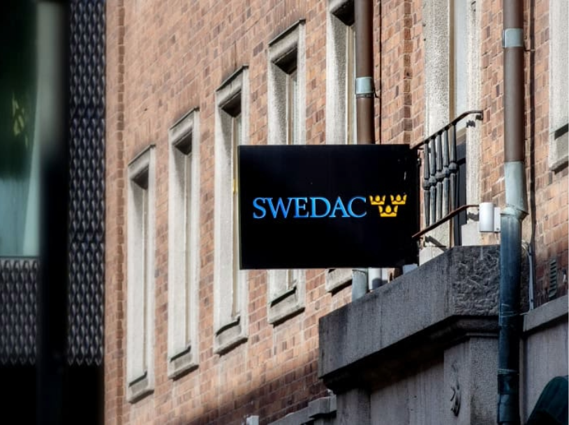 Svart skylt med texten "SWEDAC" och tre gula kronor på en tegelvägg.