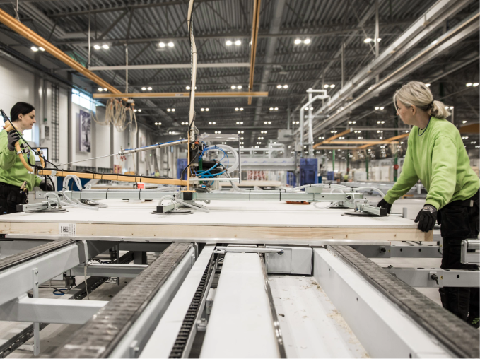 Två personer i gröna tröjor arbetar vid en maskin i en modern fabriksmiljö med industribelysning.