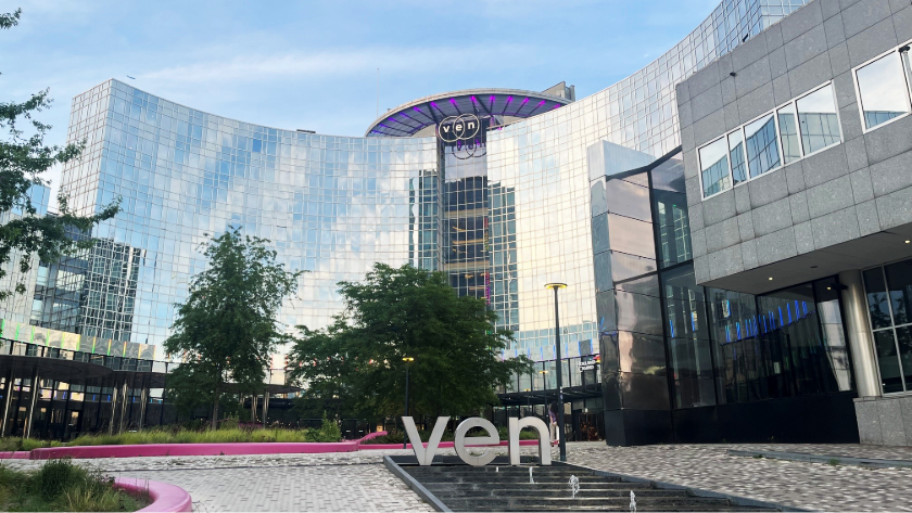 Modern glass building with "ven" sign in front, trees, cobblestone path, and reflective windows.