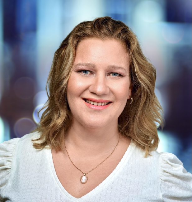 Woman with curly blonde hair smiles at the camera, wearing a white sweater and pearl necklace, blurred blue background.
